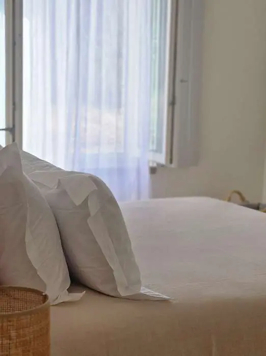 White pillows on a made bed beside a window hung with sheer linen