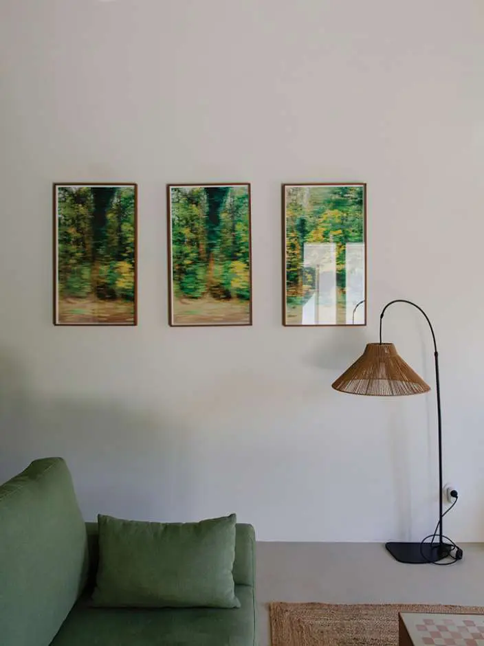 Three framed prints of trees above a green sofa, a rattan floor lamp alongside