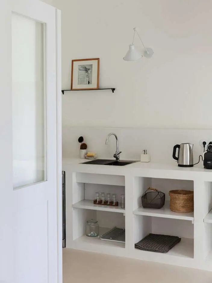 A small white kitchen with open shelves, a kettle and a framed print above the sink