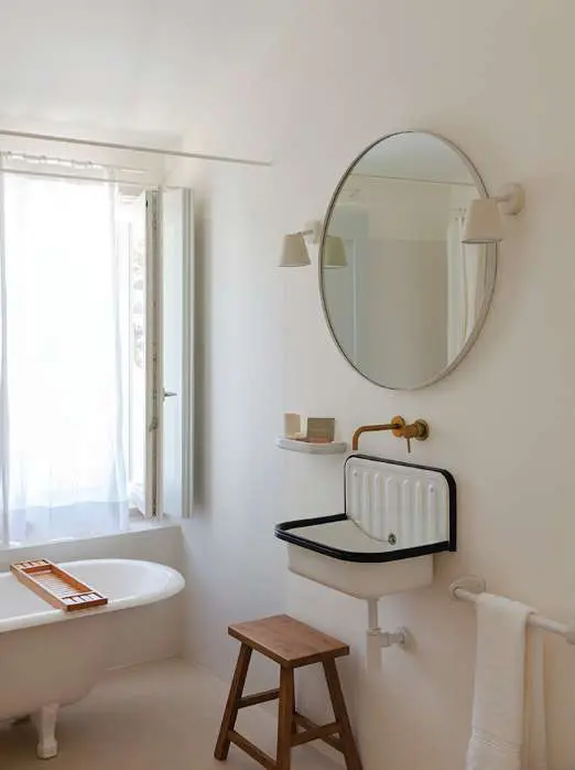 A bathroom with a roll-top bath, a round mirror and a brass tap at the window