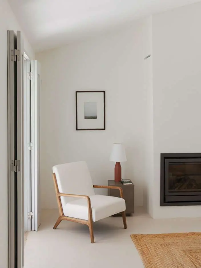 A white sitting room with a linen armchair beside a wood-burning stove